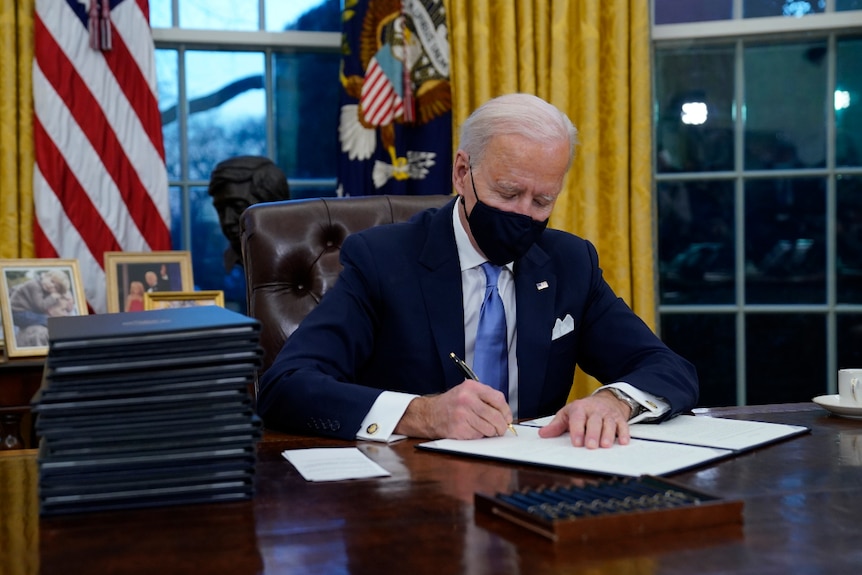 Joe Biden portant un costume et un masque facial dans le bureau ovale, signant des ordres exécutifs.