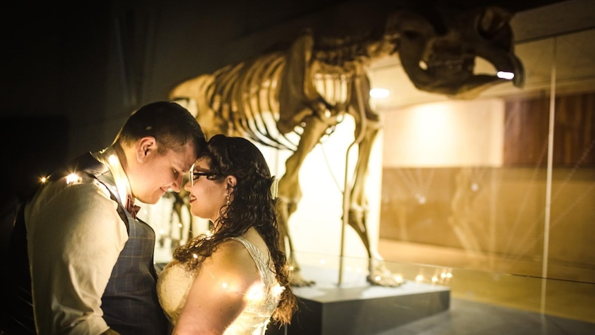 Couple dressed in wedding gown and suit in front of a dinosaur skeleton.