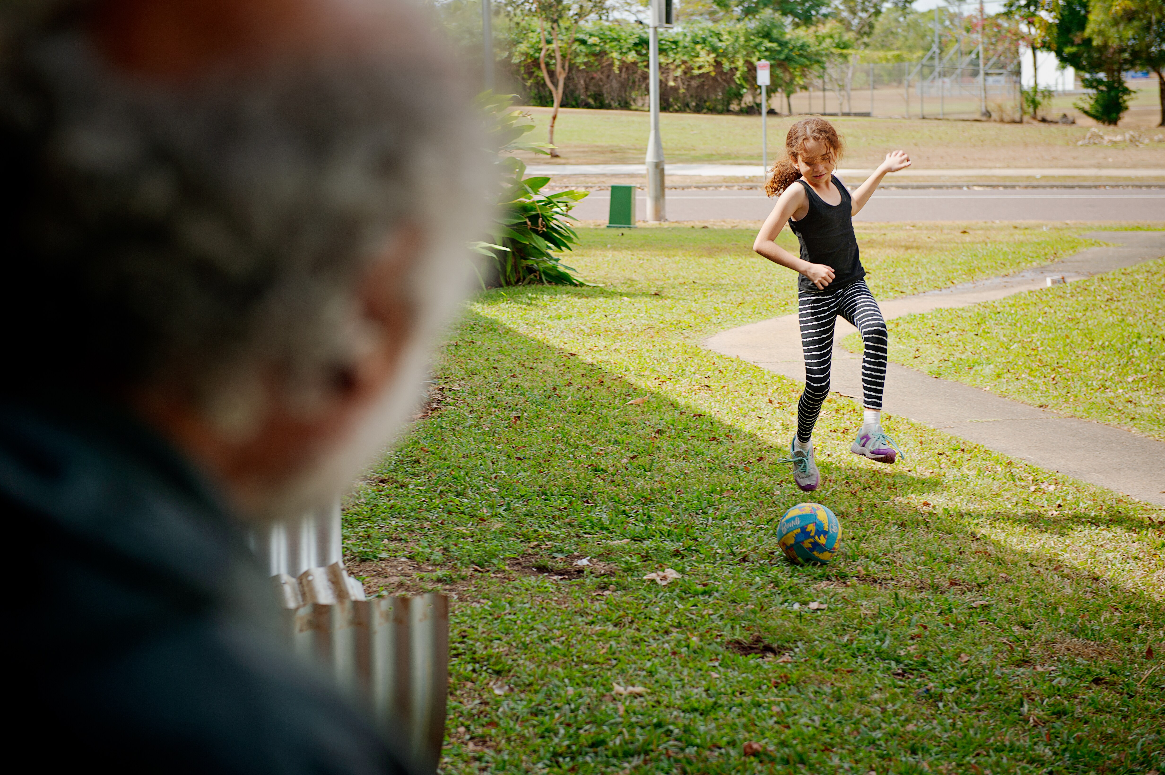A man sits on his porch and watches his young daughter kick a soccer ball on the lawn.