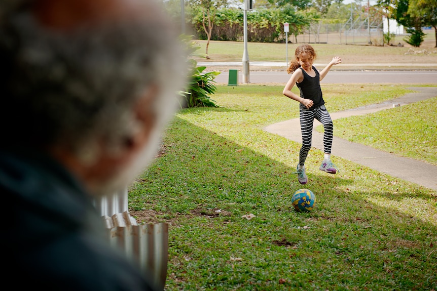 A man sits on his front porch and watches his young daughter kick a soccer ball on the lawn.