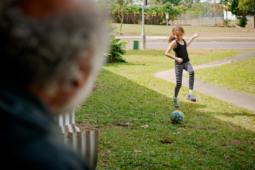 Un homme est assis sur son porche et regarde sa jeune fille frapper un ballon de football sur la pelouse.