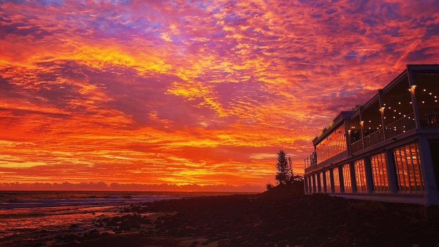 A vibrant sunrise is reflected in the windows of a two-storey building on the beach.