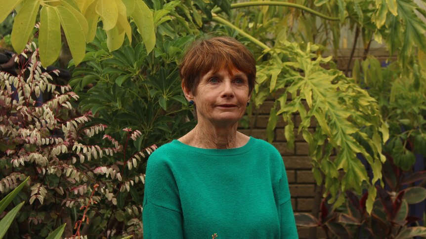 She stands in a green jumper, surrounded by green plants in a garden