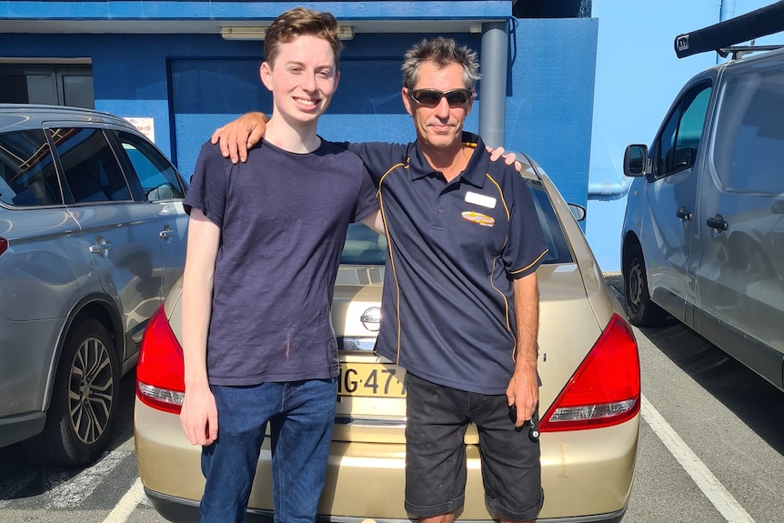 Teenage male and a man standing in front of a gold sedan with their arms over each other's shoulders 