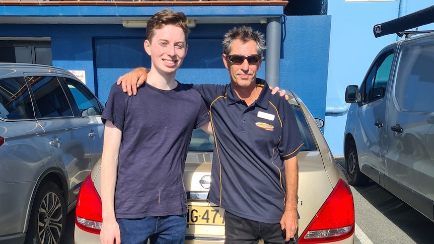 Teenage male and a man standing in front of a gold sedan with their arms over eachother's shoulders. 