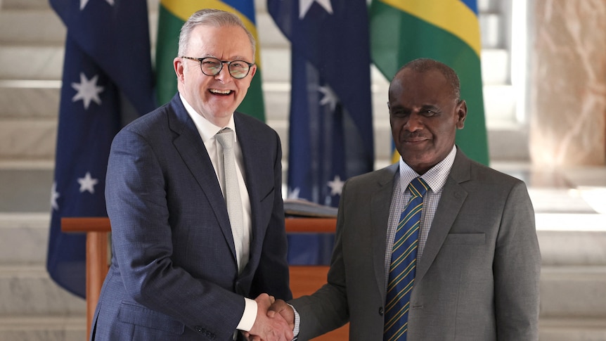 Anthony Albanese and Jeremiah Manele shake hands in front of a row of Australian and Solomon Islands flags.