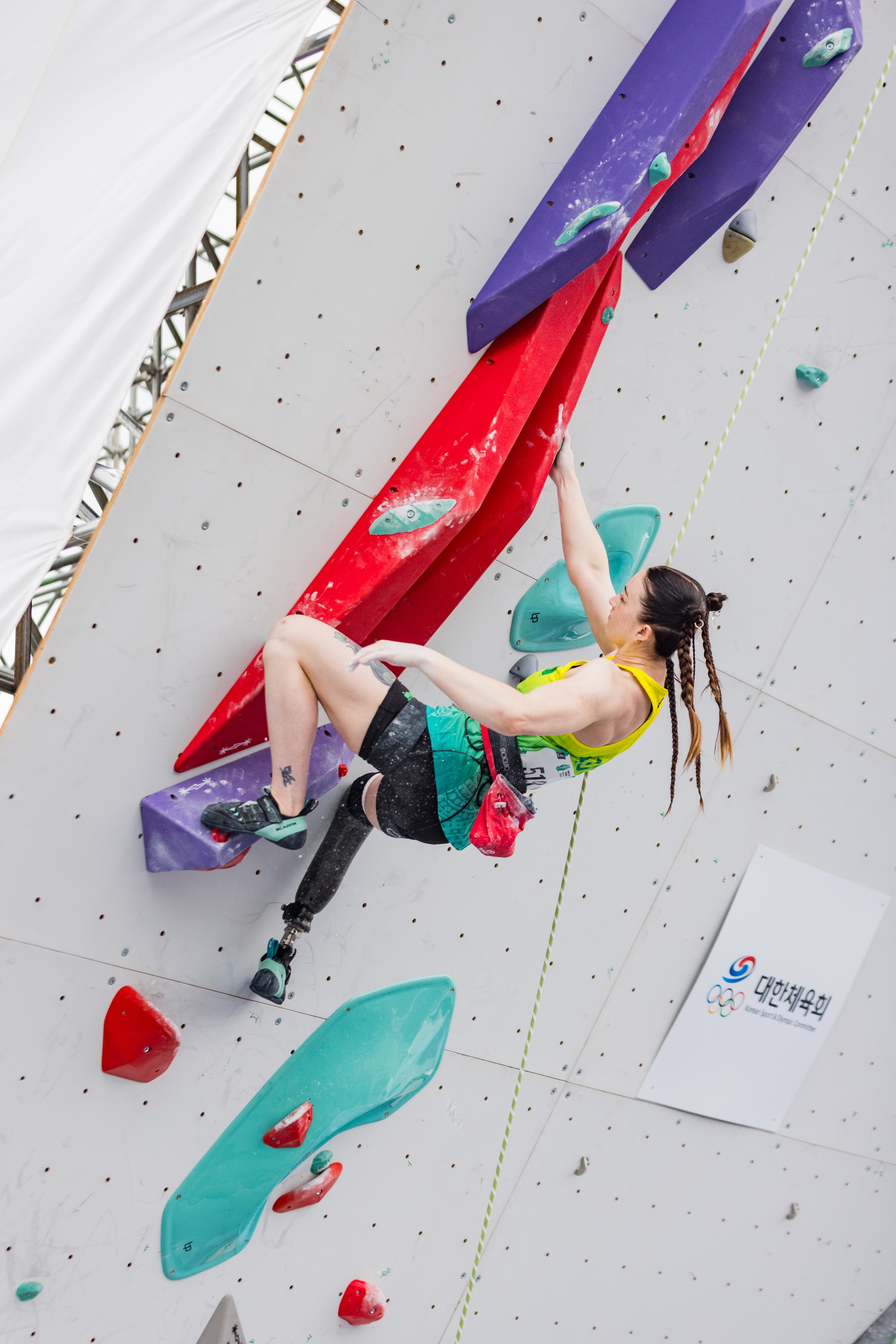 A alpinista Sarah Larcombe, usando uma camiseta dourada e tranças no cabelo, está pendurada em um suporte vermelho na parede com uma das mãos.