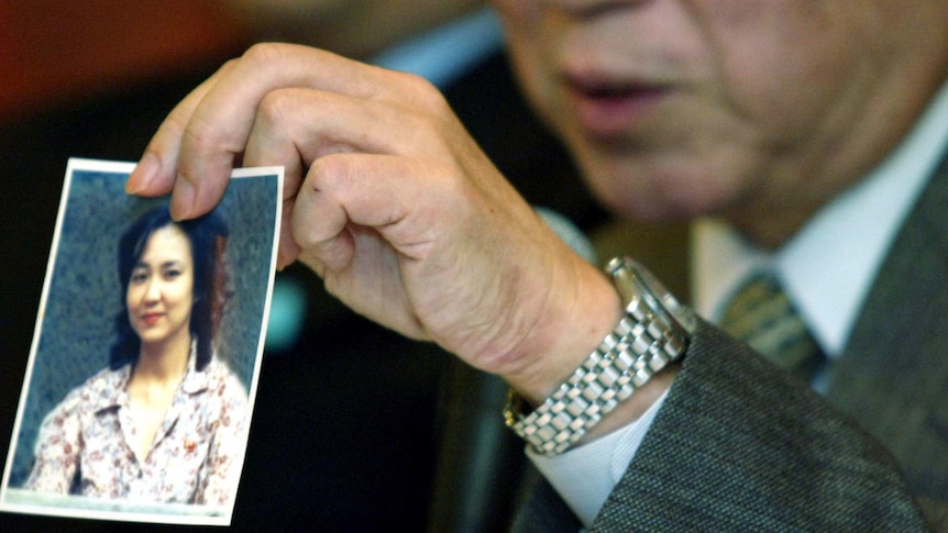 An out-of-focus elderly man in a suit holds up a photo of a young woman in a floral blouse.