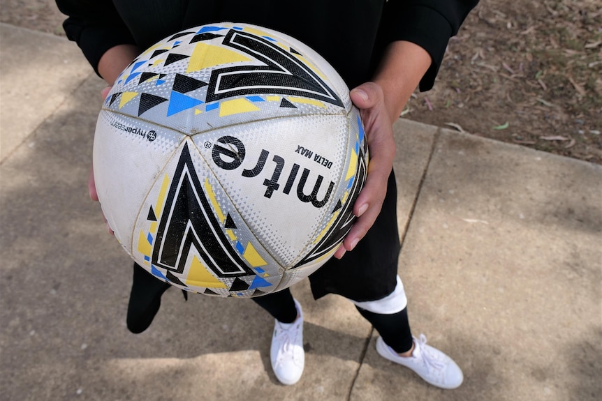 A close up of a soccer ball with a yellow, black and blue geometric pattern held by a woman wearing black leggings.