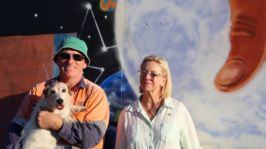 A man holding a dog next to a woman stand in front of a mural artwork.