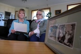 Two women sit reading an old letter in front of a black and white photo.