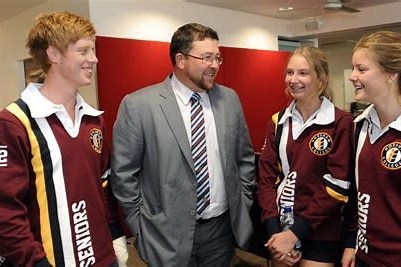 A man in a suit smiles as he speaks with three high school students in uniforms.