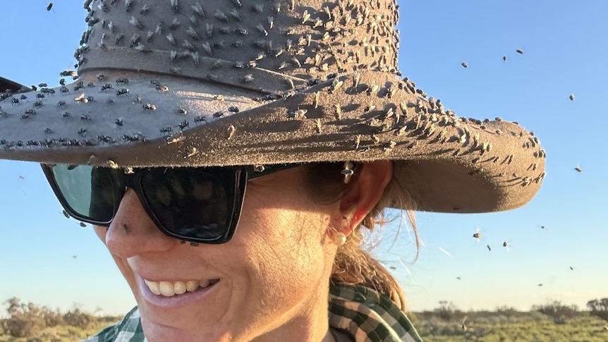 A woman in sunglasses wears a broadbrimmed hat crawling with hundreds of flies.