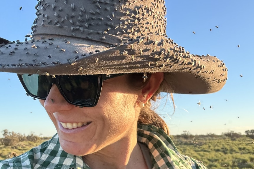A woman in sunglasses wears a broadbrimmed hat crawling with hundreds of flies.