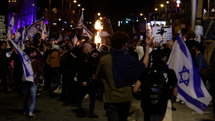 crowds gathered with israeli flag at night, fire in background