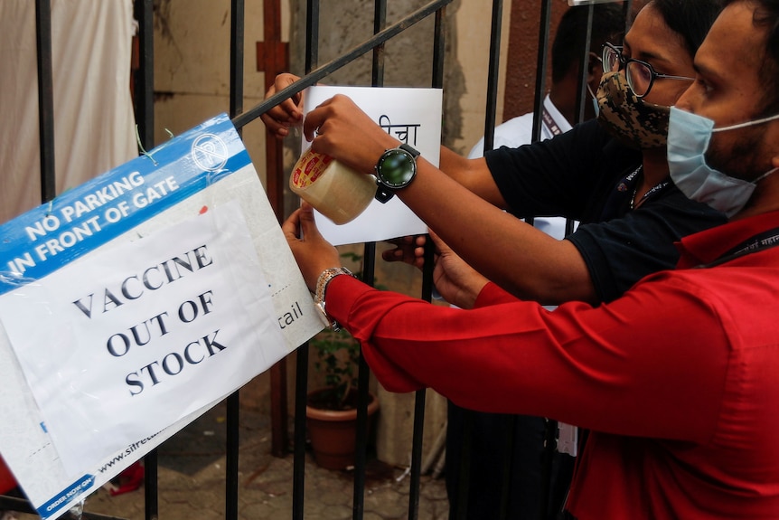 A masked man and woman tape signs to a fence saying "Vaccine out of stock".