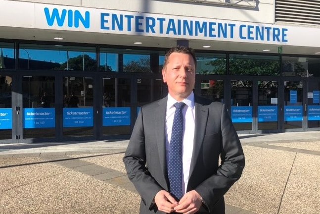 A man with dark hair, wearing a blue suit, standing outside the WIN Entertainment Centre.