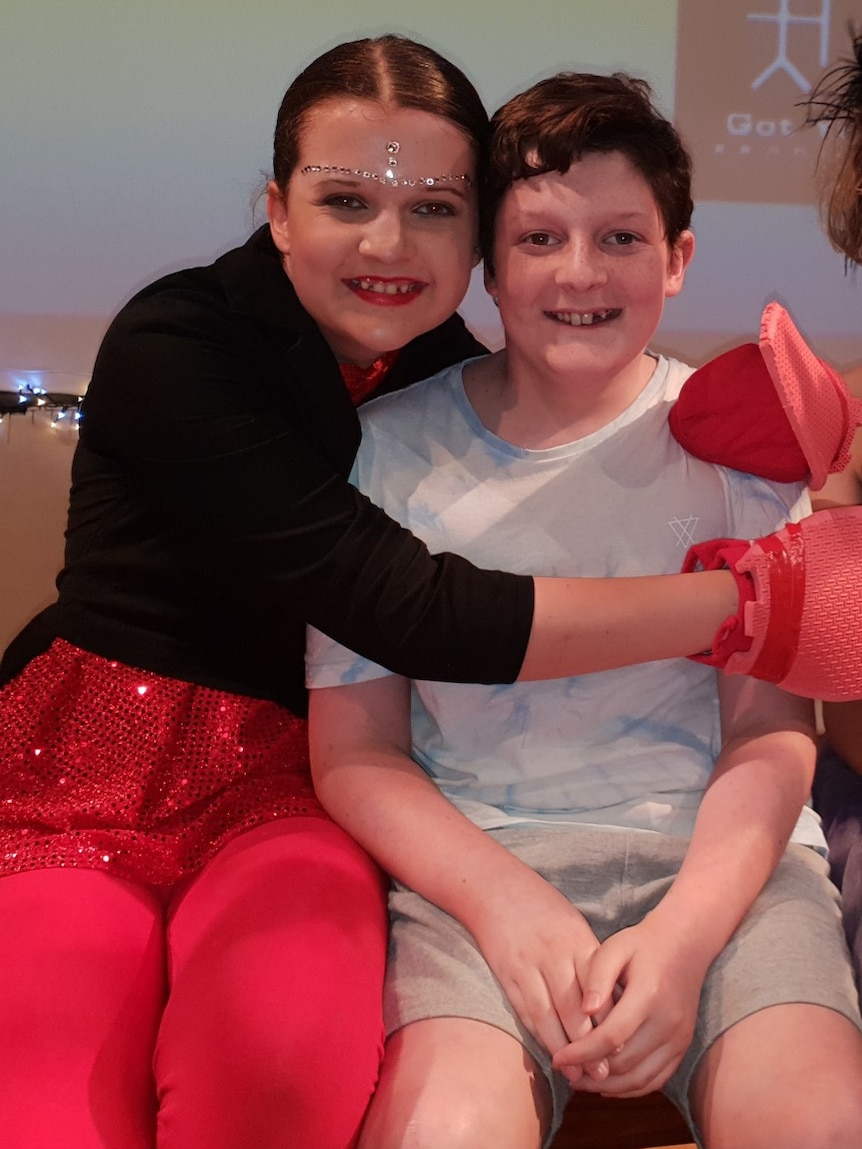A girl in a black jacket and red sequinned skirt, with sparkles on her face, hugs her brother.