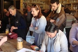 A man & women stand by the counter as another women writes her story in an A3 folio