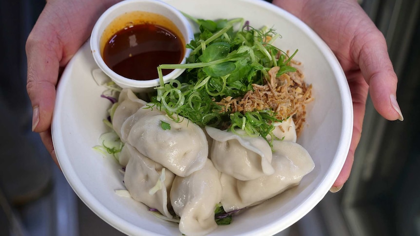 Two hands cupped around a plate of Chinese dumplings, greens and dipping sauce.