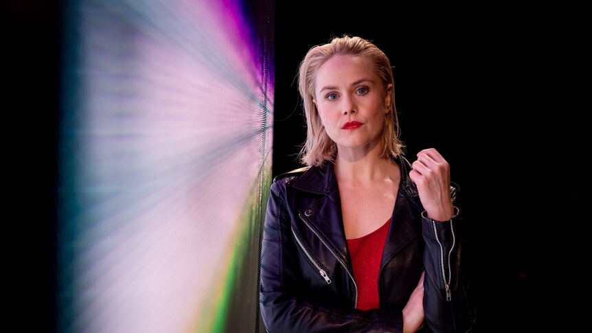 White woman with blonde lob wears red dress and leather jacket and stands with arms folded and head titled toward LED screen.