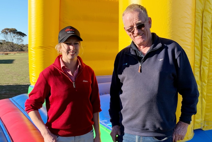 A woman with blonde hair wearing a red jumper stands next to a man wearing a navy blue jumper in front of a bouncy castle.
