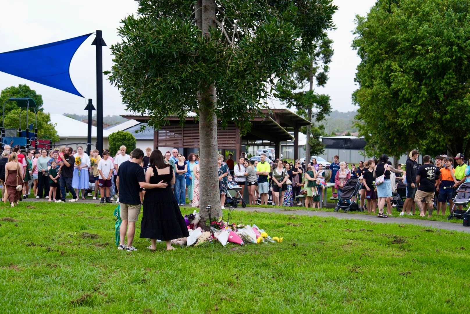 A crowd of people stand gathered in a park. Two people stand together next to a tree where people have laid flowers.