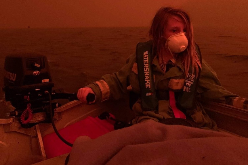 A boy in a face mask sits in a boat. Smoke and flames have made the air red and dark.