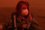 A child wears a facemask and steers a boat on the water with the red-orange sky behind them.