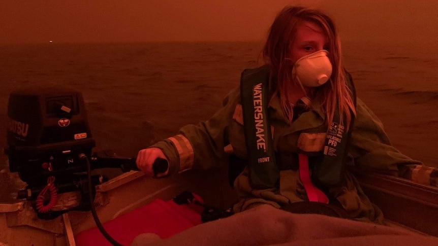 A boy in a face mask sits in a boat. Smoke and flames have made the air red and dark.