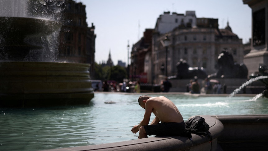 A man is sitting on the edge of a fountain leaning forward, looking defeated by the heat.