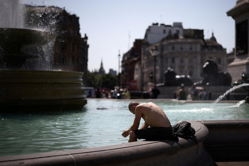 Un hombre está sentado en el borde de una fuente inclinado hacia adelante, luciendo derrotado por el calor.