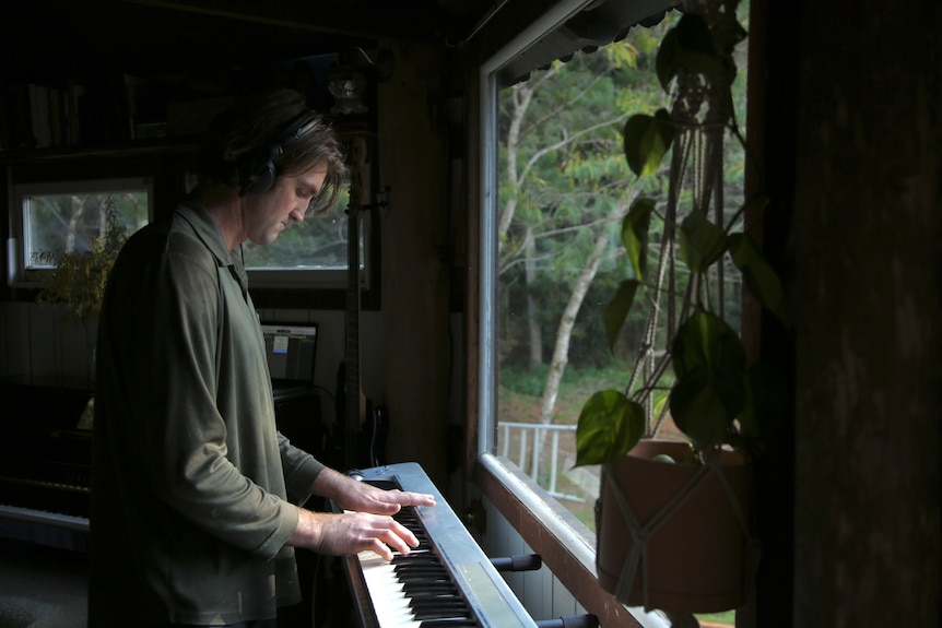 A man stands playing the keyboard in front of a large window. Greenery can be seen outside