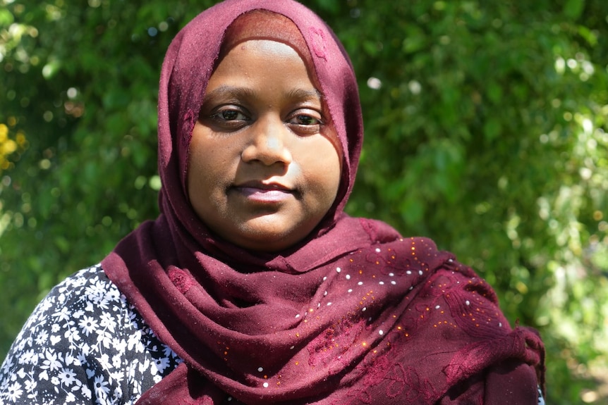 A woman in a maroon hijab with a slight smile looks at the camera, leaves and greenery behind.