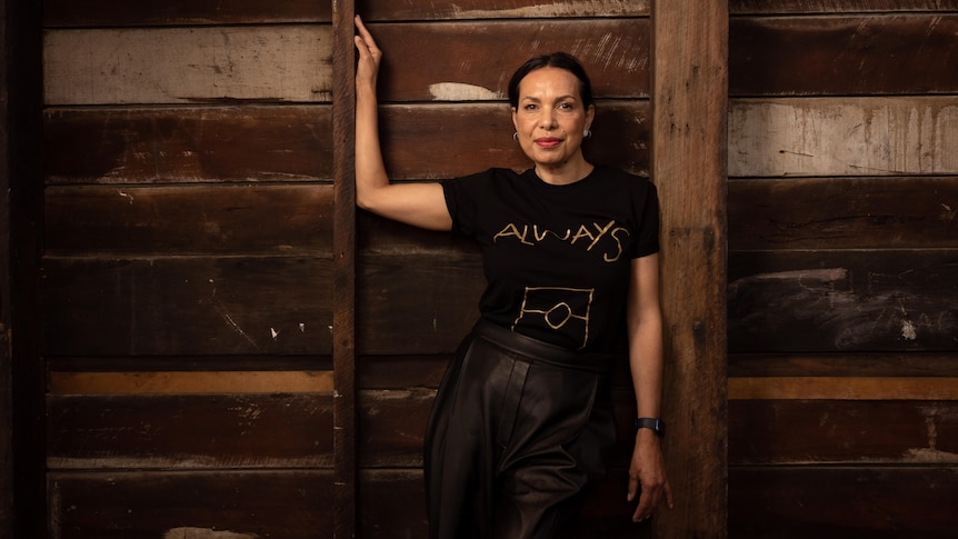 A Kokatha woman with black hair pulled back wears a black shirt that reads 'Always' in gold and stands against wooden wall