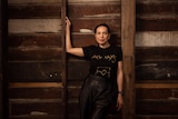 A Kokatha woman with black hair pulled back wears a black shirt that reads 'Always' in gold and stands against wooden wall