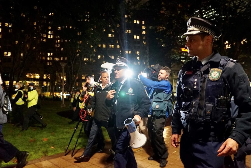 Police and journalists walk through a park holding a loudspeaker.