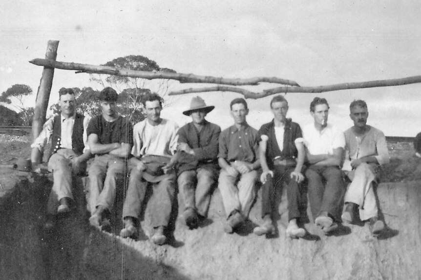 A historic photo of eight men sitting side by side on top of an irrigation trench.