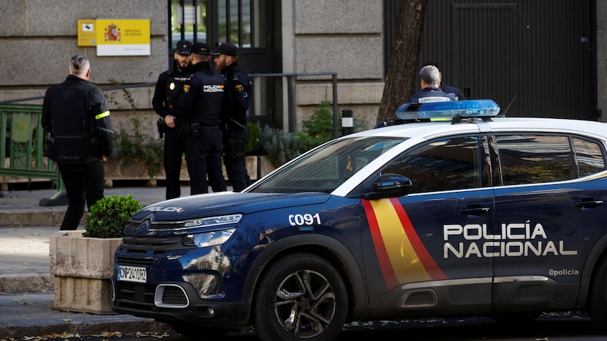 Police officers stand outside Spain's Ministry of Defence, with police car parked on curb.
