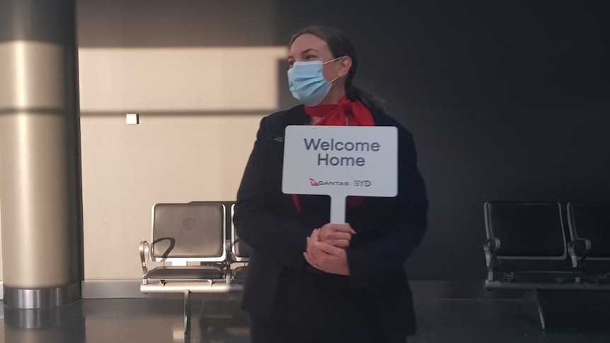 A masked woman stands in an airport terminal holding a sign that reads "Welcome home".