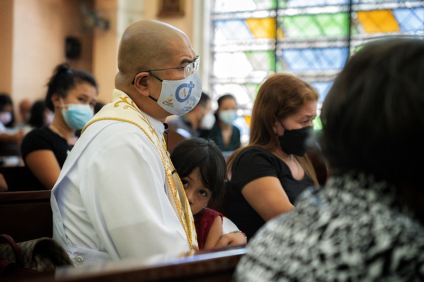 A Filipino priest in a mask holds a young girl to his side in a pew 