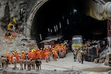 Dozens of natual disaster personell, dressed in orange gather around a tunnel, waiting for trapped workers to be rescued