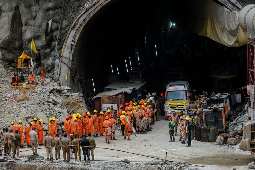 Dozens of natual disaster personell, dressed in orange gather around a tunnel, waiting for trapped workers to be rescued