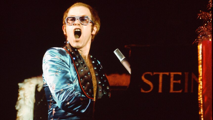 Elton John performs live at a piano during 1973 London show, mouth open at the audience
