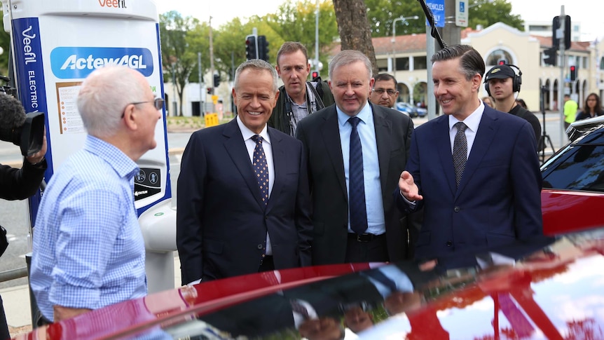 The three men smile and look at a red electric car parked in front of them