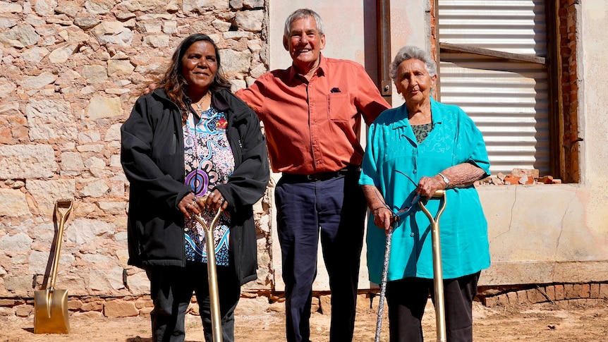 Two indigenous women standing either side of a man with an orange shirt and holding golden shovels.