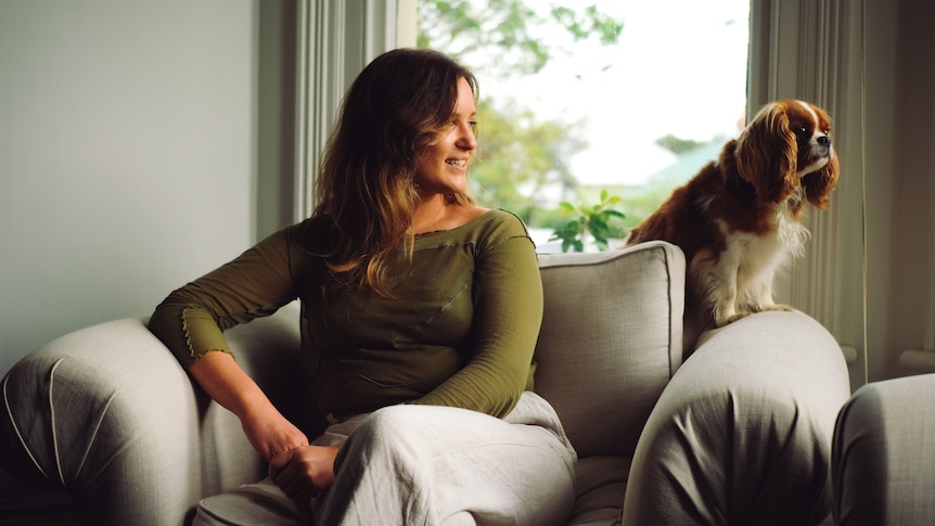 A blonde woman sits on a grey sofa with a cavalier king charles to her right as she looks right. She wears a green top.