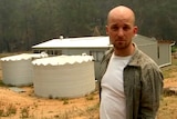 A man stands on a rural property in front of a house and water tanks.