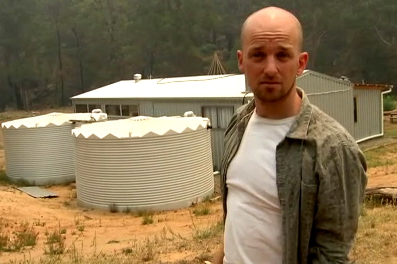A man stands on a rural property in front of a house and water tanks.