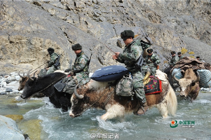 A group of Chinese soldiers ride yaks through a clea, rocky mountain stream.